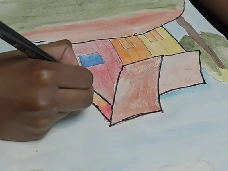 Detailed view of a hand outlining a colorful house drawing with a black pen.