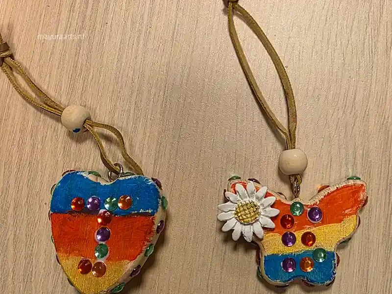 Two handmade craft pendants on leather cords, featuring a heart and a butterfly shape painted in rainbow colors and decorated with sparkling sequins.
